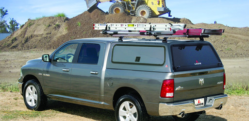 A gray pickup truck with a covered bed and ladders on a roof rack is parked on a dirt field. In the background, a large yellow excavator is moving soil. Trees and a clear sky are visible.