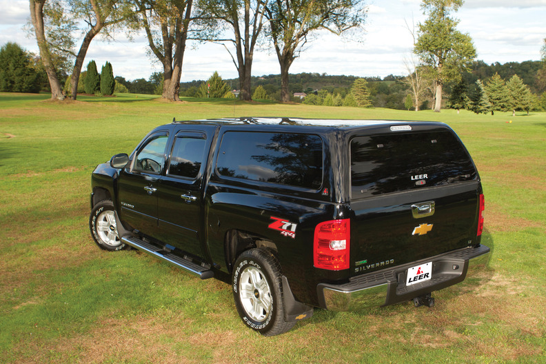 A dark green pickup truck with a canopy is parked on grass in a rural area. Trees and a small body of water are visible in the background under a clear blue sky.
