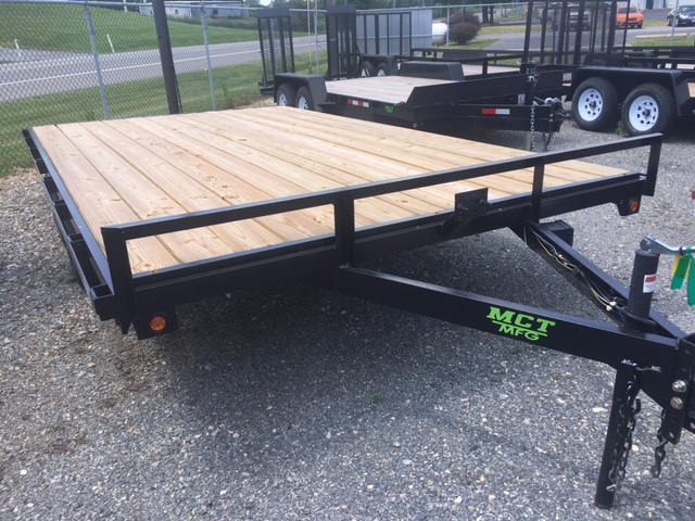 A flatbed trailer with a wooden deck and black metal frame is parked on a gravel surface. It has a hitch in the front and is situated near a chain-link fence. Another trailer is visible in the background.