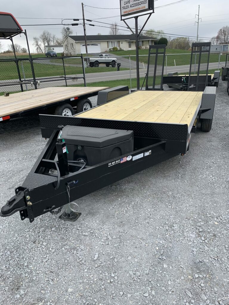 A flatbed trailer with a wooden deck and metal frame is parked on gravel. The trailer has a toolbox mounted on the front and ramps at the back. Nearby are other trailers and a fence, with buildings and a street in the background.