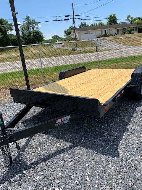 A flatbed trailer with a wooden deck is parked on a gravel lot. It has a metal frame and a hitch for towing. In the background, there's a chain-link fence, a road, and a few houses with trees and a clear blue sky.