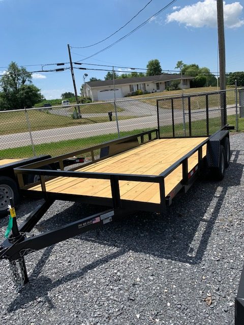 A flatbed utility trailer with a wooden platform and metal frame is parked on gravel. It is hitched to the tow bar, with a chain link fence and a road in the background under a clear sky.