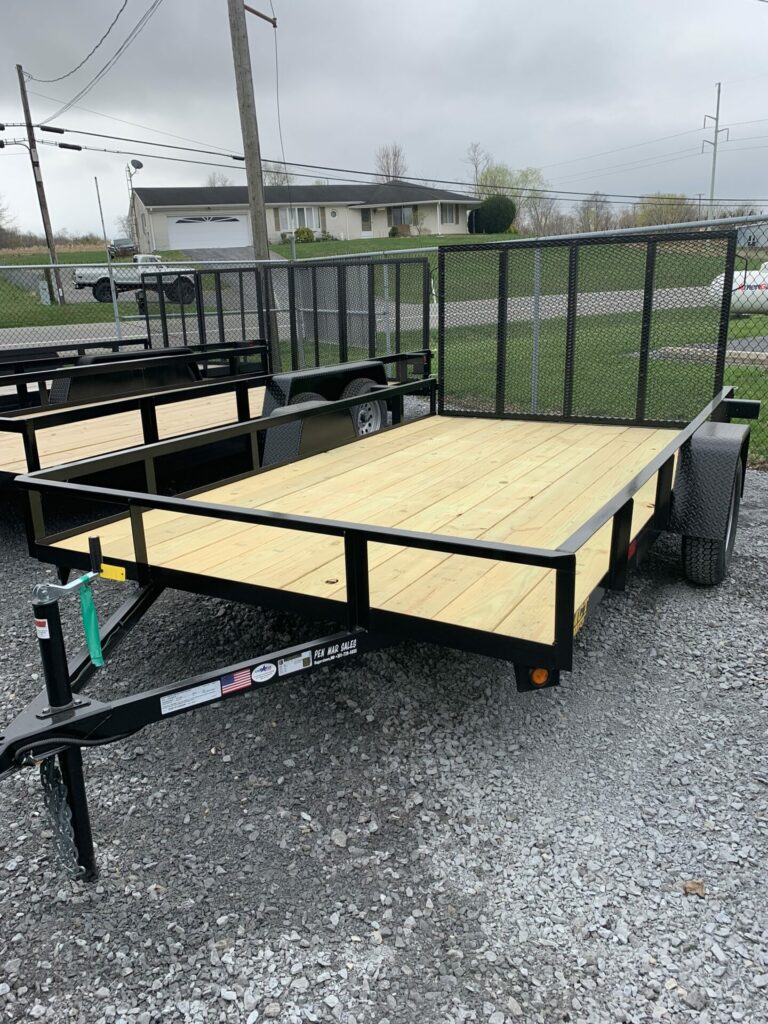 A flatbed utility trailer with a wooden deck and metal frame is parked on a gravel surface. It features a mesh ramp gate at the back and has single axle with black wheels. A house and some trees are visible in the background under a cloudy sky.