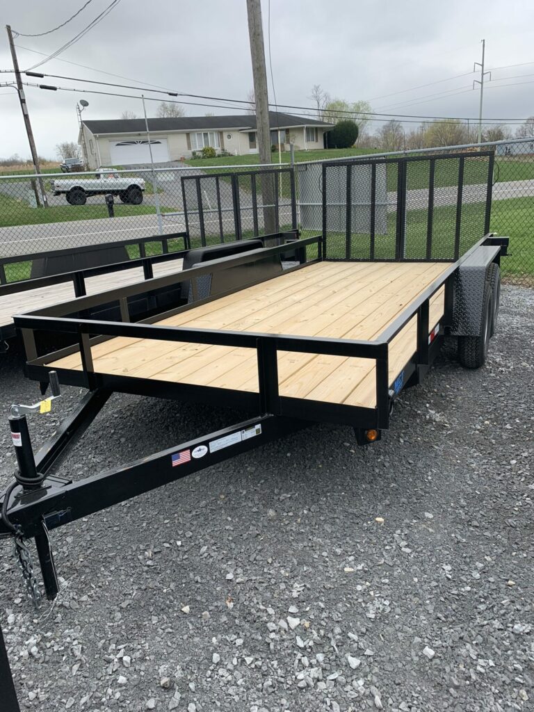 A flatbed utility trailer with a wooden deck and metal frame is parked on gravel. It has a railing on three sides and a single axle. In the background, there is a chain-link fence, a house, and trees under an overcast sky.