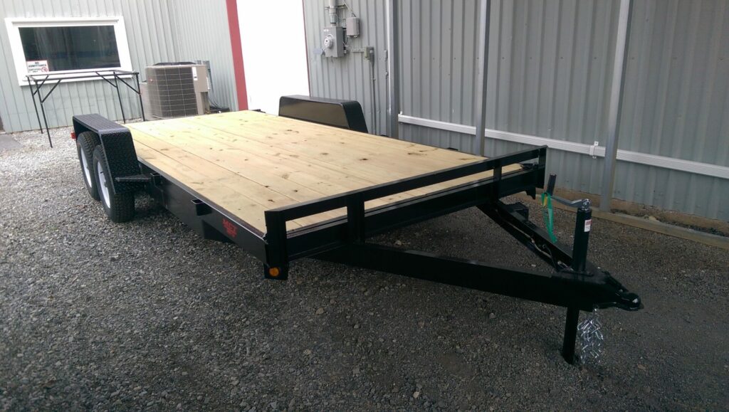 A flatbed trailer with a black metal frame and a wooden bed, parked on gravel next to a metal building. It has two wheels on each side and a hitch for towing at the front. An air conditioning unit is visible in the background.