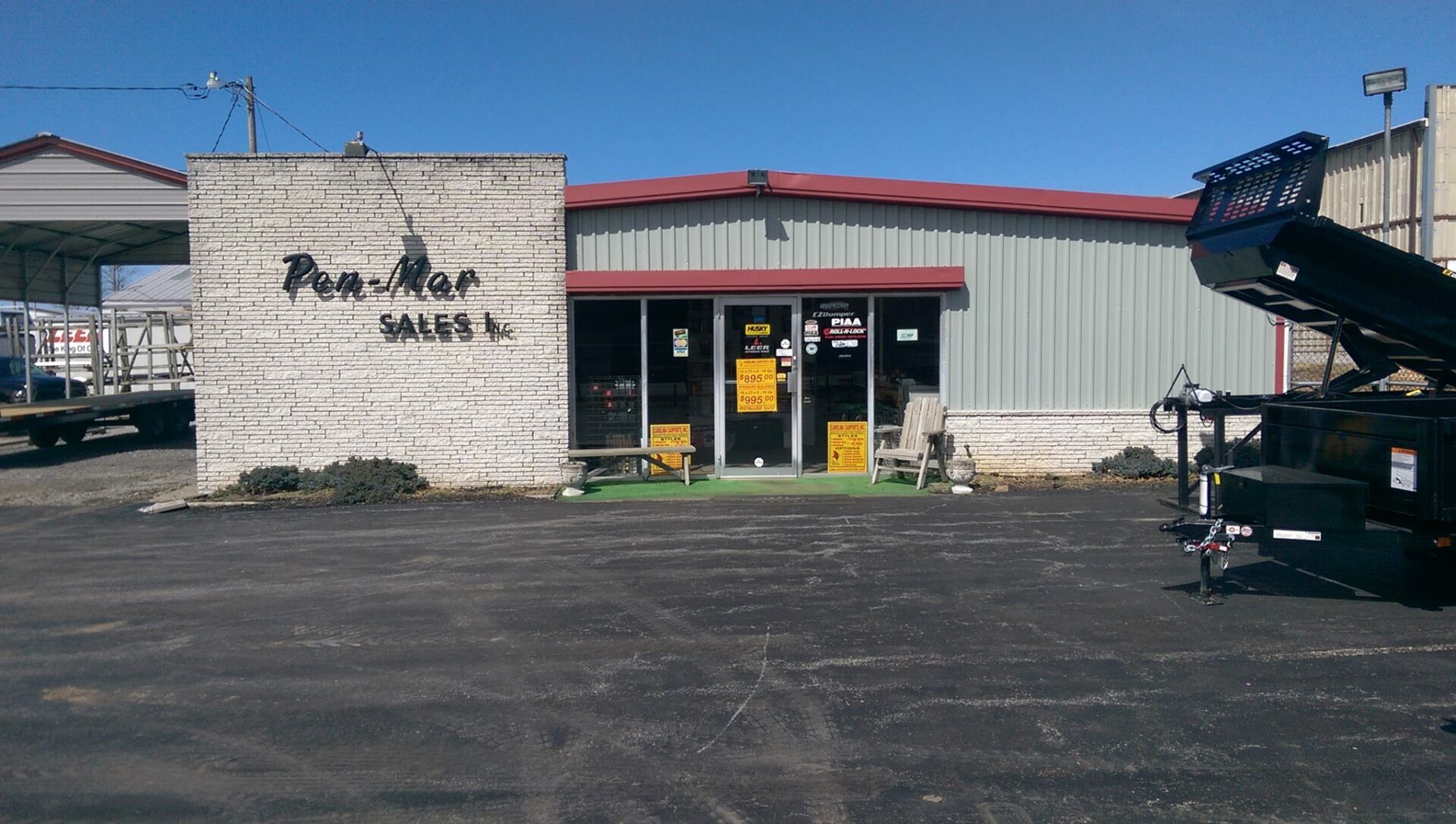 Exterior view of a building with a sign reading "Pen Mar Sales." The entrance is glass with several notices posted. A large piece of equipment is parked nearby. The sky is clear and blue.