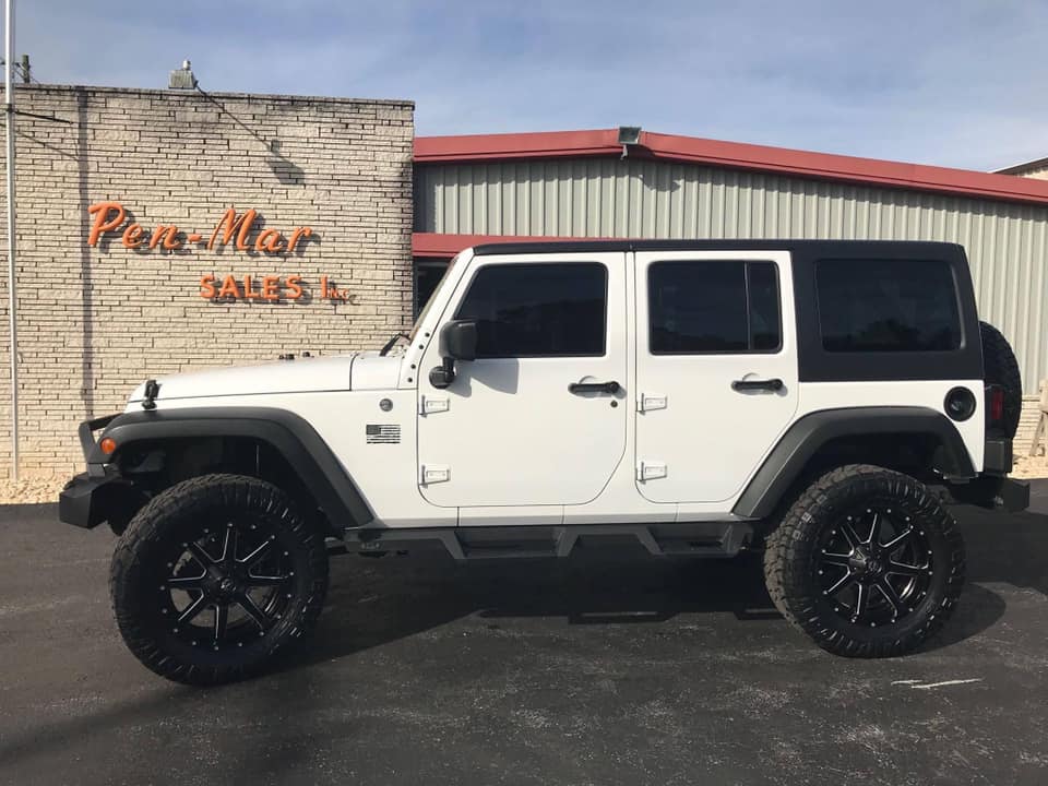 A white, four-door Jeep with large black rims is parked in front of a building with a sign reading Pen-Mar Sales. The building has a brick facade and a garage door. The sky is clear.