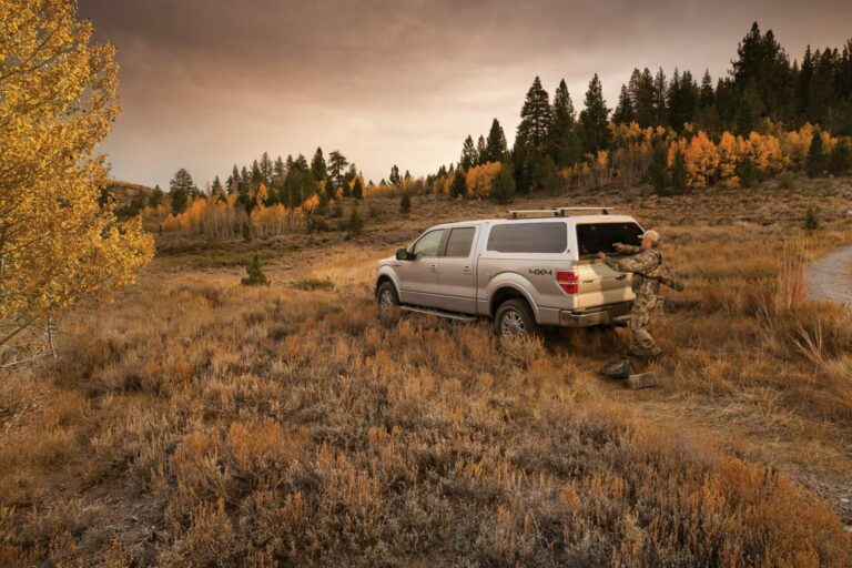 A person in camouflage unloads equipment from a white SUV parked off-road in a scenic autumn landscape. Yellow and orange foliage, dry grass, and coniferous trees surround them under a cloudy sky.