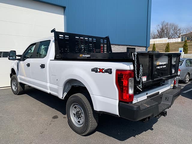 A white pickup truck with a 4x4 logo on the rear side sits parked. It features a black flatbed rack with a tailgate. The truck is parked on a paved surface near a building with a large garage door. Trees and a fence are visible in the background.