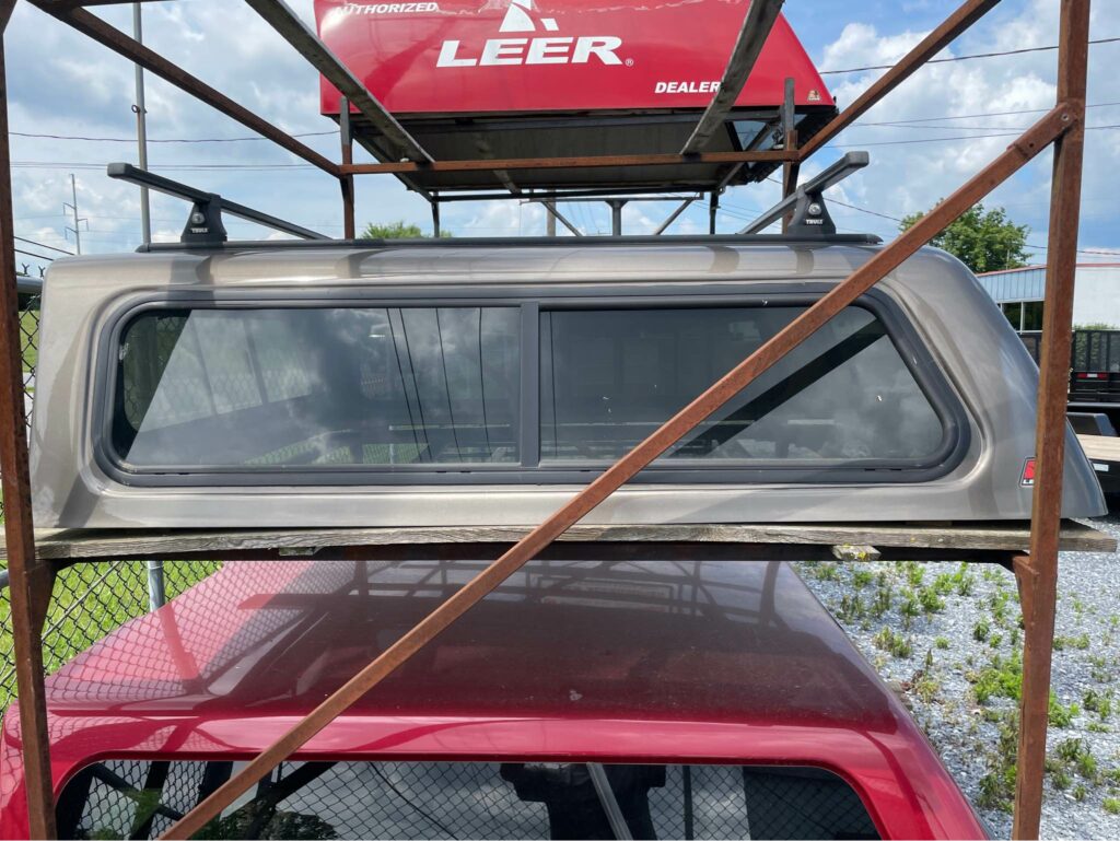 A truck cap is displayed on a metal rack outdoors. Its positioned above a red vehicle and has tinted windows. A LEER sign is visible in the background. The setting features a fence and grassy area under a partly cloudy sky.