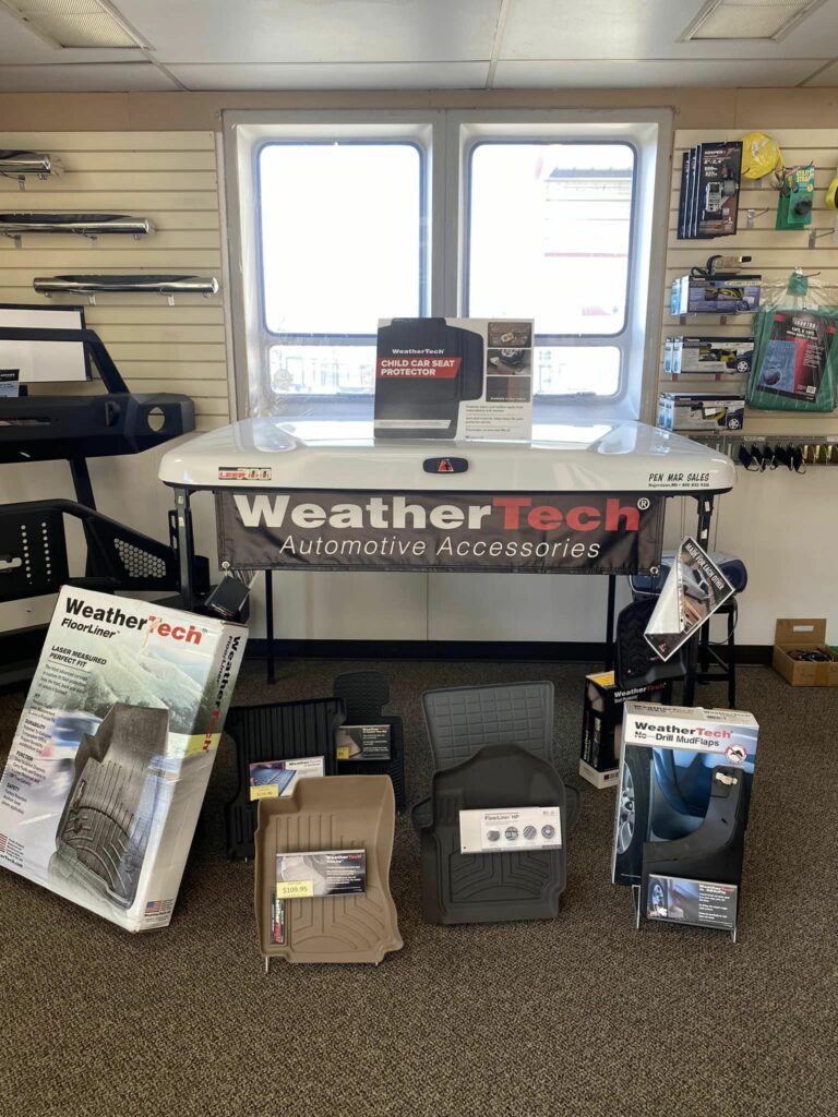 Display of WeatherTech automotive accessories inside a store. Products include floor mats, protectors, and organizers. Items are positioned around a central table with a branded banner. Shelves hold additional car-related items. Sunlight streams from a window.