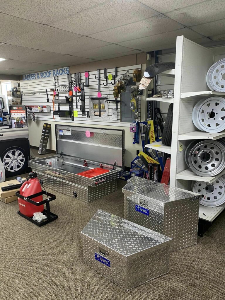 A store interior displaying automotive accessories. Visible are metallic storage boxes, a red gas can, various tools, shelves with wheel rims, and racks with roof racks and ladders. The carpet is beige, and the ceiling is made of white tiles.