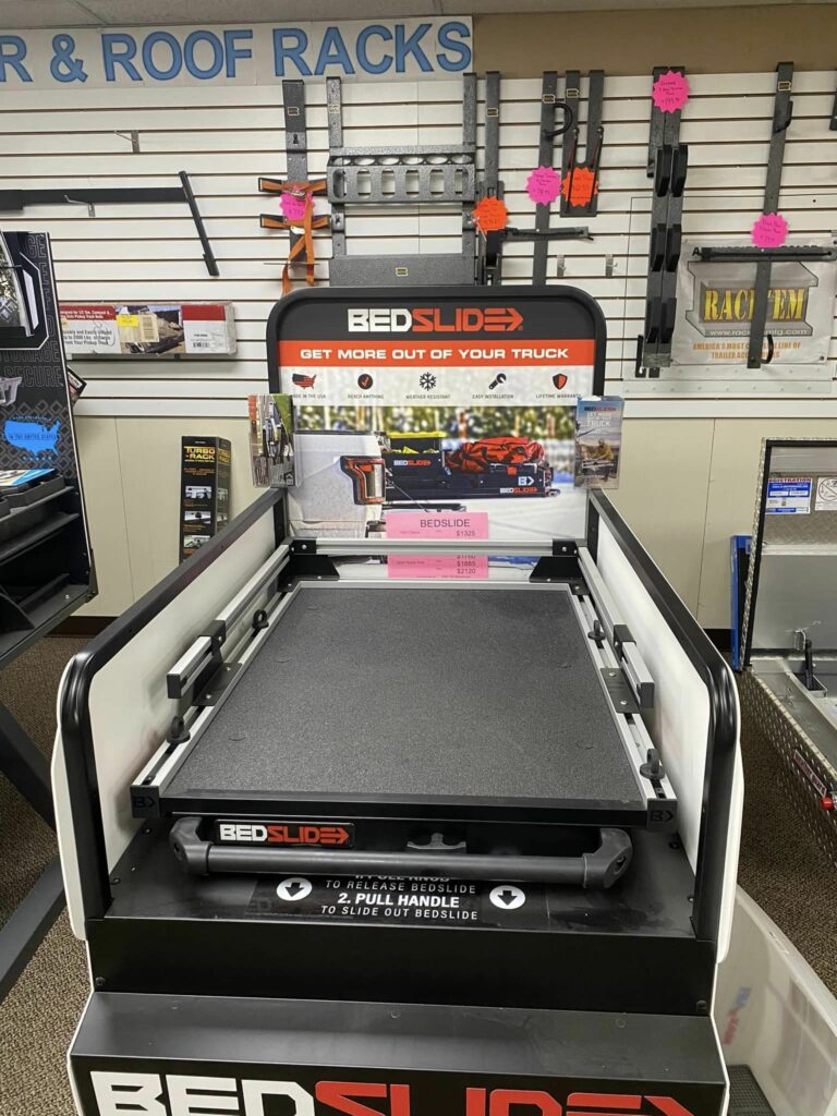 Display of a Bedslide system in a store. The device is shown extended and is surrounded by various wall-mounted tools and vibrant orange price tags. The tagline reads, Get More Out of Your Truck.