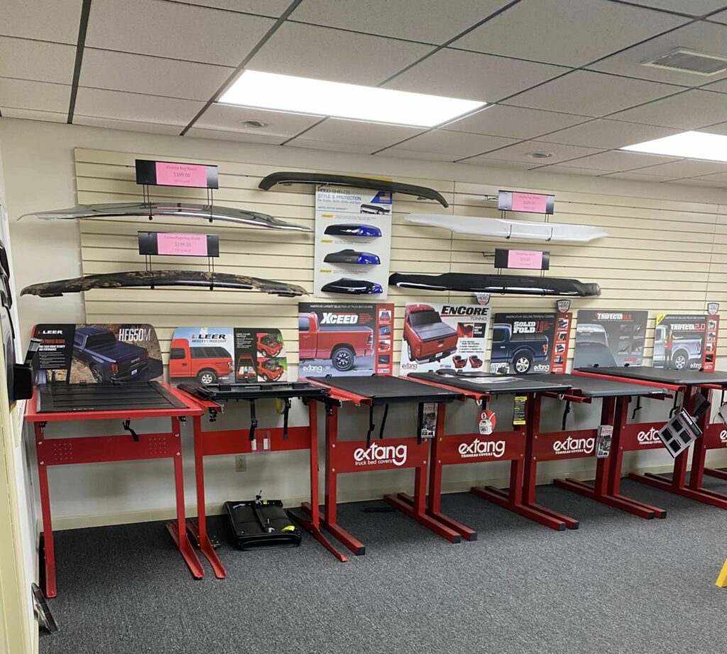 A store display shows various truck bed covers and accessories by Extang. The wall features mounted samples and boxes, with a row of red workstations underneath. Ceiling lights illuminate the gray carpeted floor.