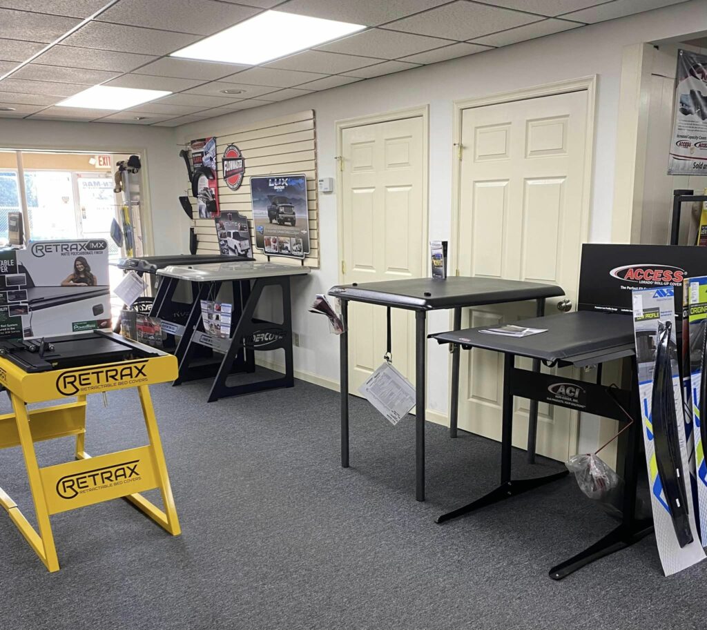 Store interior displaying various truck bed covers on stands. The room has gray carpet and white walls with promotional materials. Two doors are at the back, and sunlight filters through a window by the entrance.