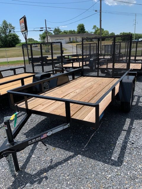 A flatbed utility trailer with a wooden floor and metal frame is parked on gravel. It has a mesh ramp at the back, spare tire, and a hitch. Several similar trailers are visible in the background under a clear sky.