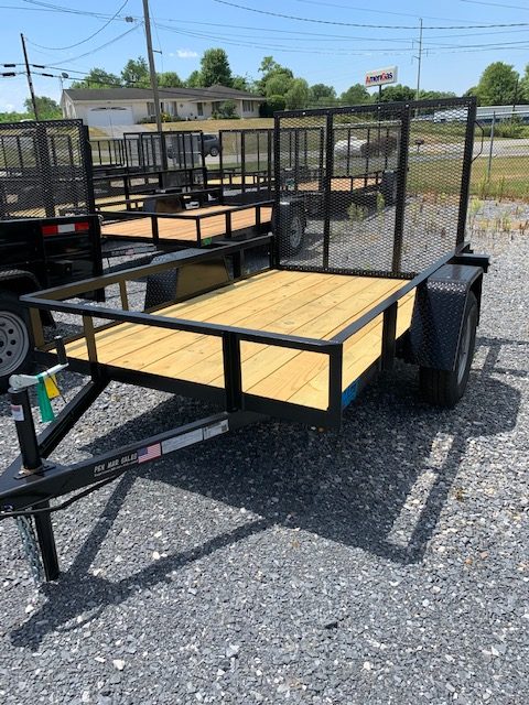 Utility trailer with a wooden deck and metal mesh sides on display in a gravel lot. Other trailers and a building with greenery are visible in the background.