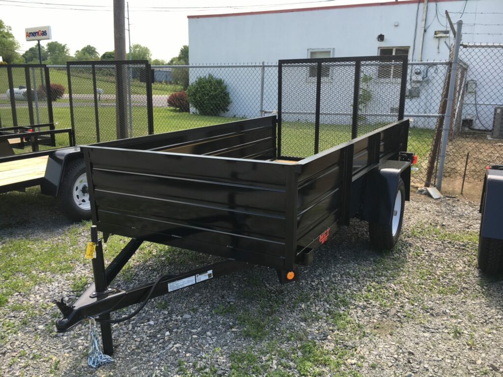 Open black utility trailer with metal sides and a rectangular frame parked on gravel near a chain-link fence. A building with a white exterior is in the background, surrounded by green grass and shrubs.