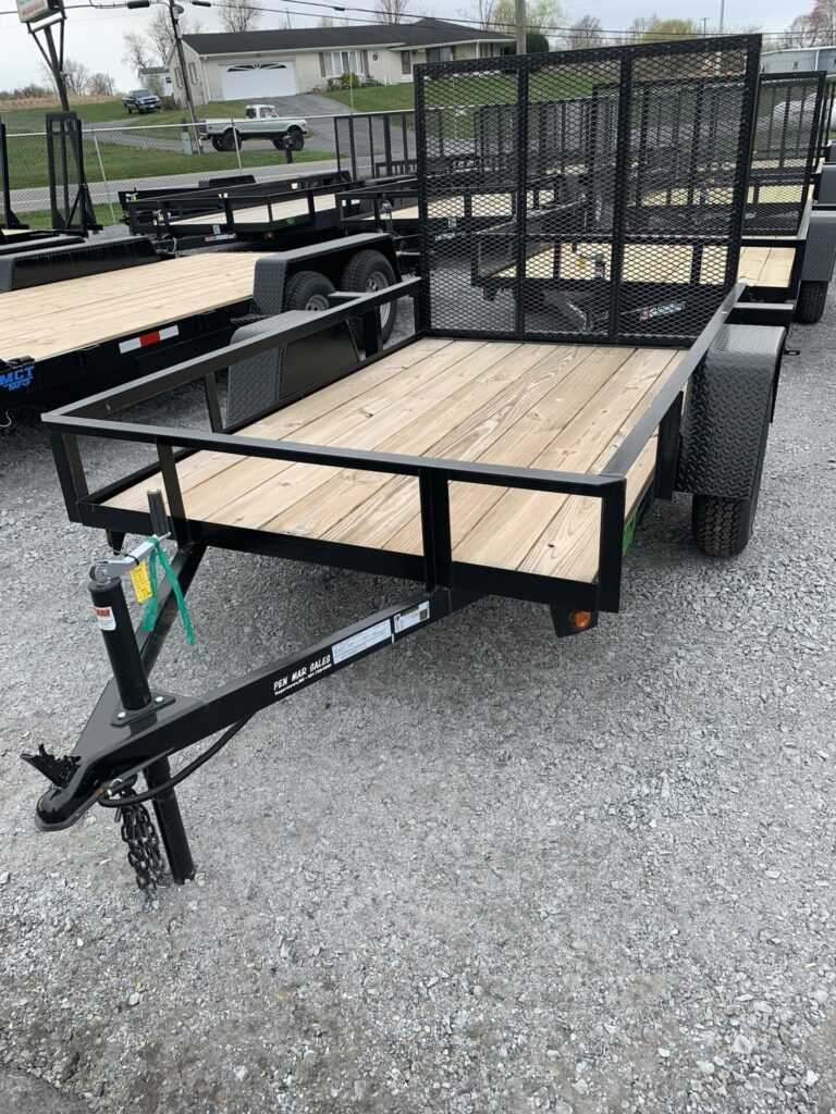 A flatbed utility trailer with a wooden deck and metal frame is parked on a gravel lot. It features a mesh ramp gate at the back, a hitch at the front, and sturdy wheels on each side. Other similar trailers are visible in the background.