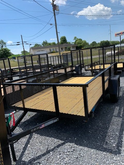 A display of multiple flatbed trailers with wire mesh sides and wooden floors, parked on gravel under a clear blue sky. Power lines and a building are visible in the background.