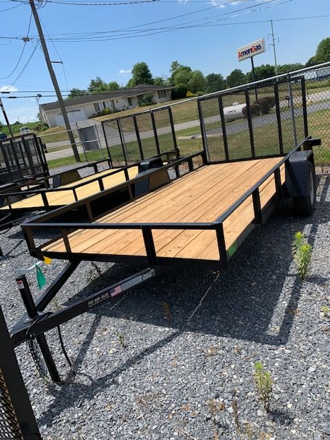 A flatbed utility trailer with wooden flooring is parked on a gravel surface near a chain-link fence. In the background, there's a grassy area, a road, and a building with a sign reading "AmeriGas." Small weeds are visible on the ground.