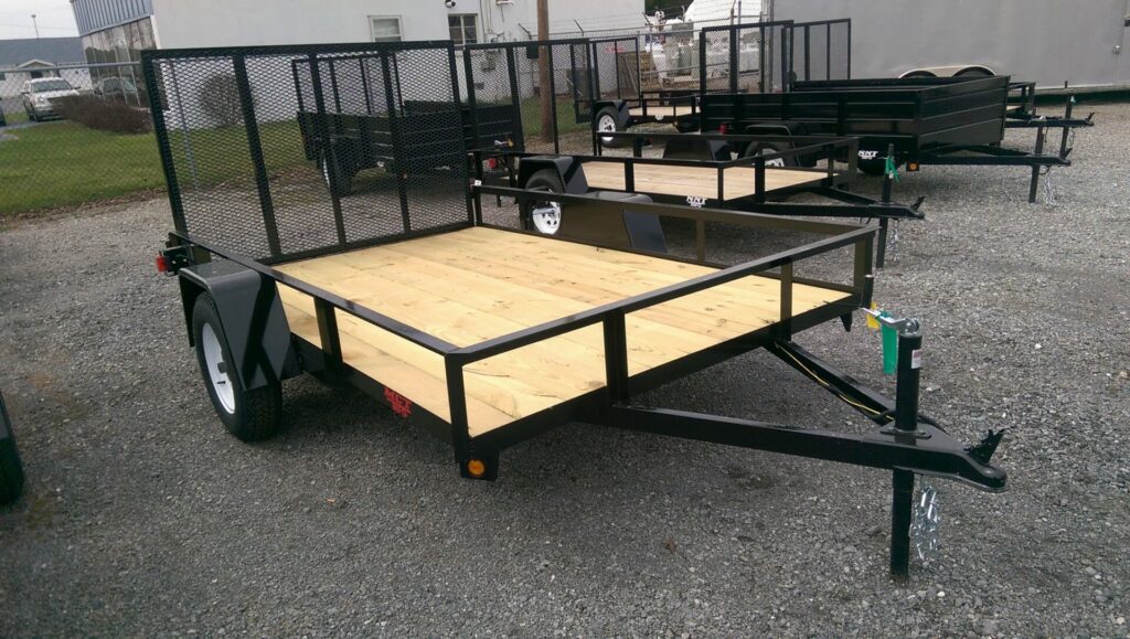 A flatbed utility trailer with a wooden deck and a mesh metal ramp at the back is parked on gravel. Other similar trailers are visible in the background, along with a chain-link fence and buildings.