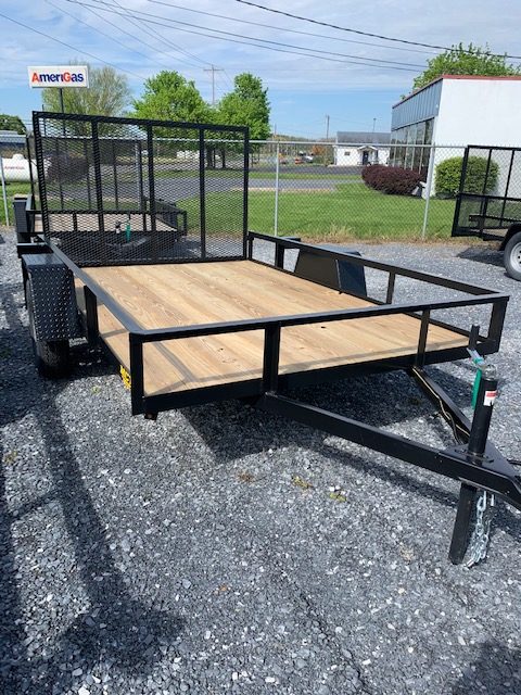 A flatbed utility trailer with a wooden deck and black metal frame is parked on gravel. It has mesh sides and is located near a chain-link fence and buildings. A sign reading "AmeriGas" is visible in the background.