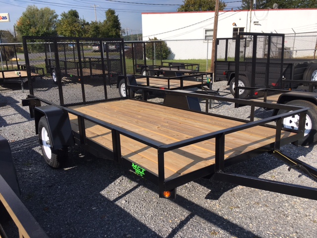 A flatbed utility trailer with a wooden deck and black metal frame, parked on gravel. It has white wheels and mesh side panels. In the background, other trailers and a white building are visible.