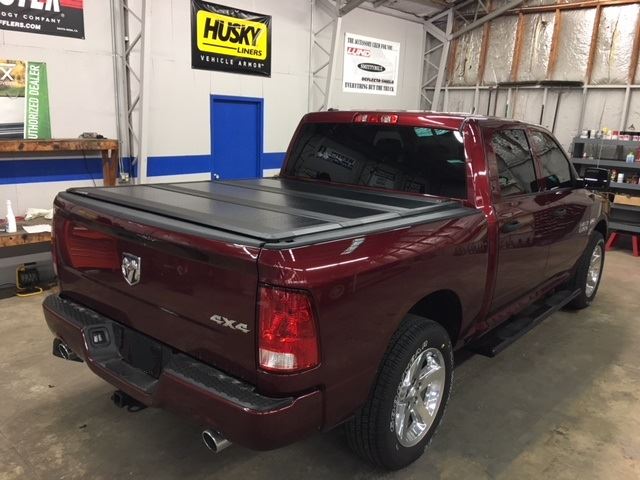 A red pickup truck with a covered bed is parked inside a garage. The truck features chrome wheels and dual exhaust. The garage walls display various automotive brand signs.