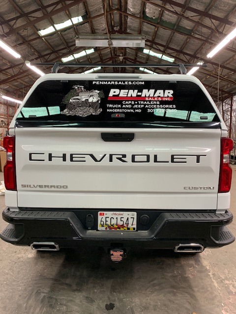 Rear view of a white Chevrolet Silverado with the word CHEVROLET on the tailgate. The rear window displays an advertisement for Pen-Mar sales. Maryland license plate 6EC1547 is visible. The scene is inside a warehouse or garage.