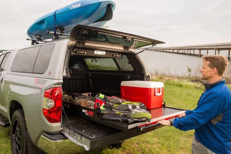 A person adjusts a sliding gear tray from the back of a silver truck, which is parked near a body of water. The tray holds a red cooler, boots, and gear. A blue kayak is secured on the roof. There is a bridge in the background.