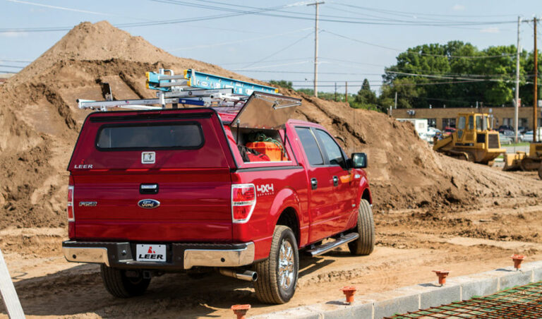 A red Ford truck with a Leer canopy is parked at a construction site. It has a ladder on the roof and construction materials inside. In the background, there are large dirt piles, machinery, and trees.