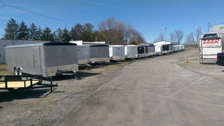 A row of silver trailers is parked on a gravel lot under a clear blue sky. Trees and a few buildings are visible in the background. A portion of a larger vehicle with the word 