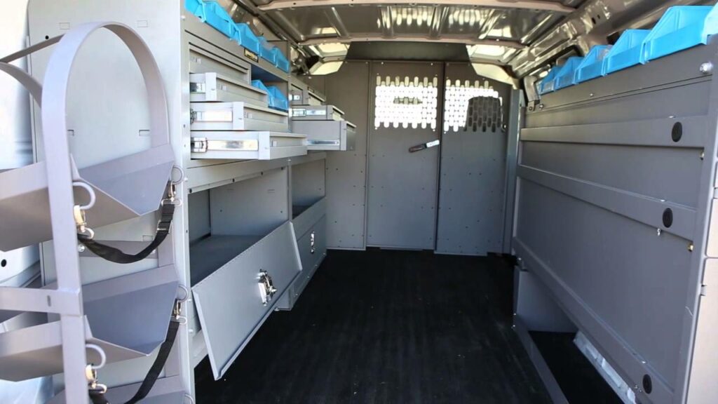 Interior of a utility van with organized shelves and compartments. The left side features blue organizers and metal drawers. The right side has adjustable shelves. The floor is black, and the back has a partition with a barred section.