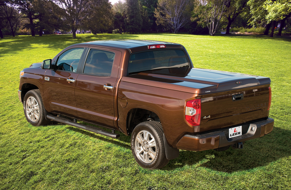 A brown pickup truck with a Leer topper is parked on green grass with trees in the background. The truck is viewed from the back, showcasing its sleek design and accessories under a clear sky.