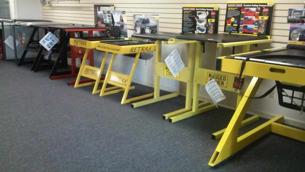 A display of workbenches in a store, featuring different models in yellow and red. Each bench is equipped with various tool storage options, and product packaging is visible on top of some benches. The floor is carpeted in gray.