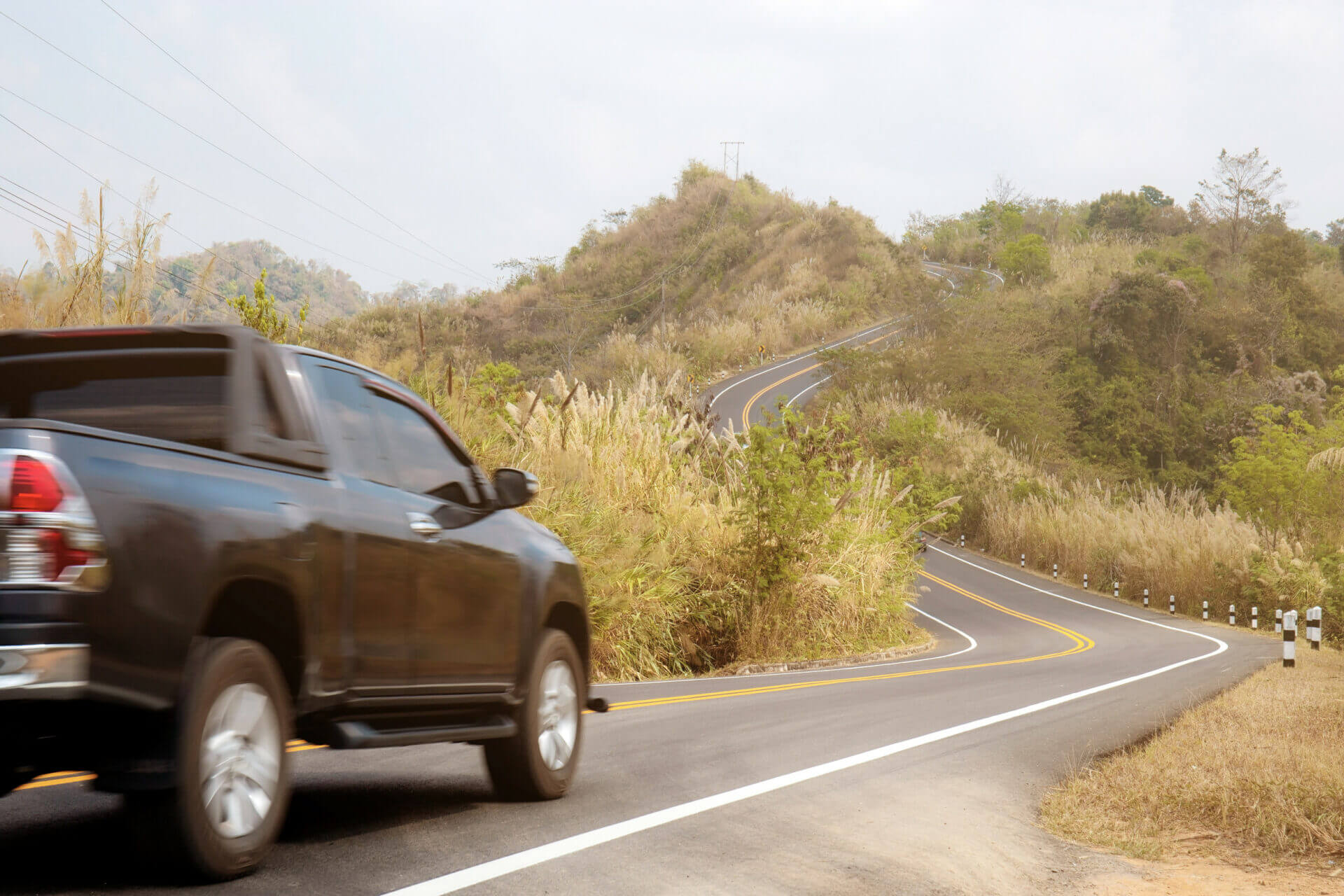 A black pickup truck drives along a winding rural road with hills and vegetation in the background.